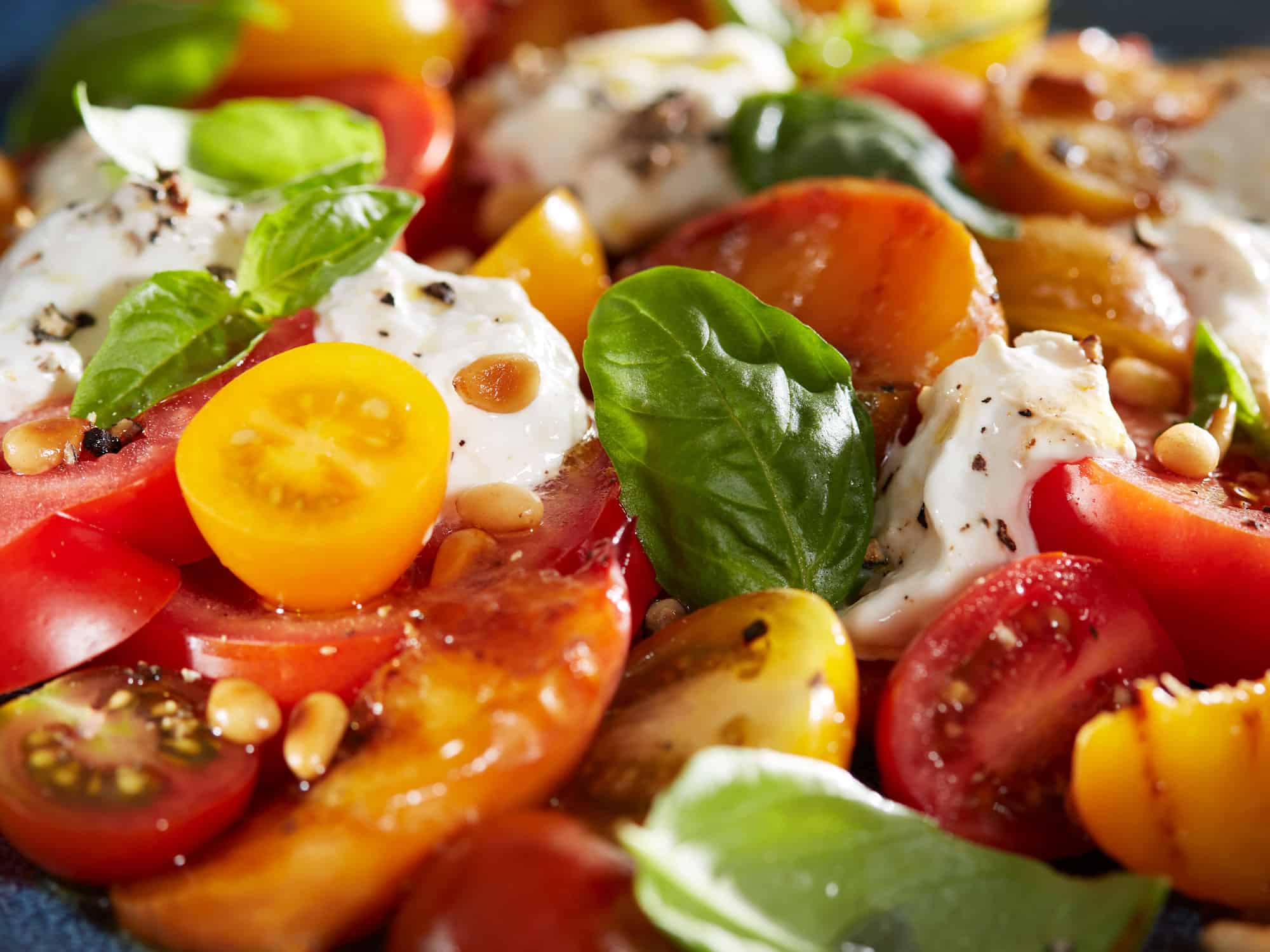 Nahaufnahme eines Salates mit bunten Tomaten, Burrata, gegrillten Pfirsichen, Basilikum und gerösteten Pinienkernen.
