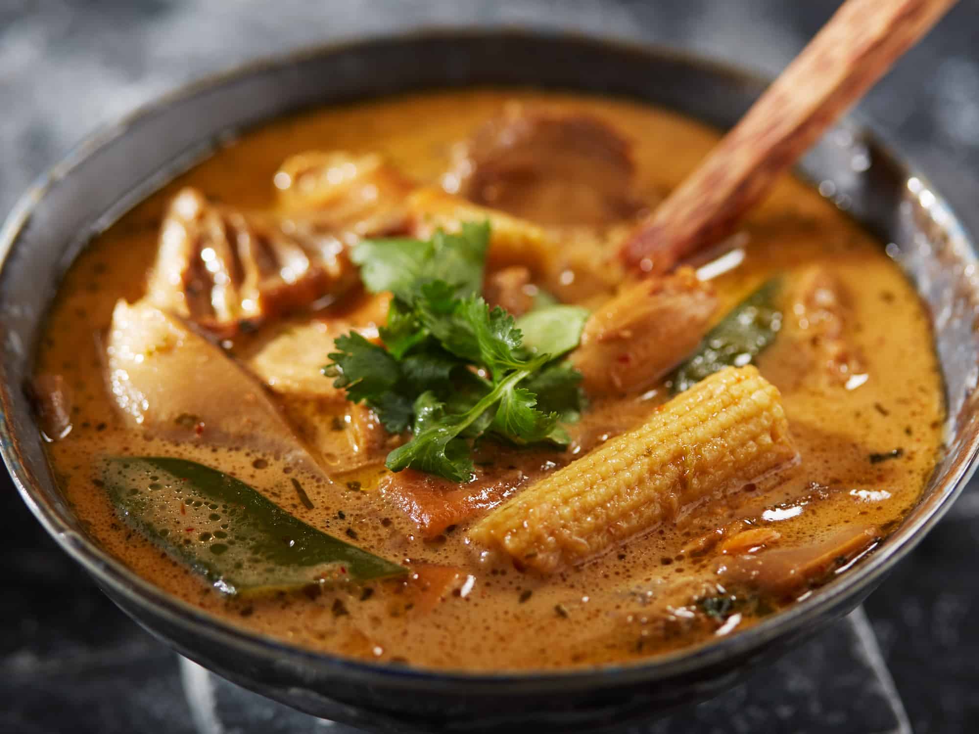 Nahaufnahme einer thailändischen Tom Kha Gai Suppe mit Hühnchen, Austernpilzen, Zuckerschoten und Maiskölbchen, garniert mit frischem Koriander.