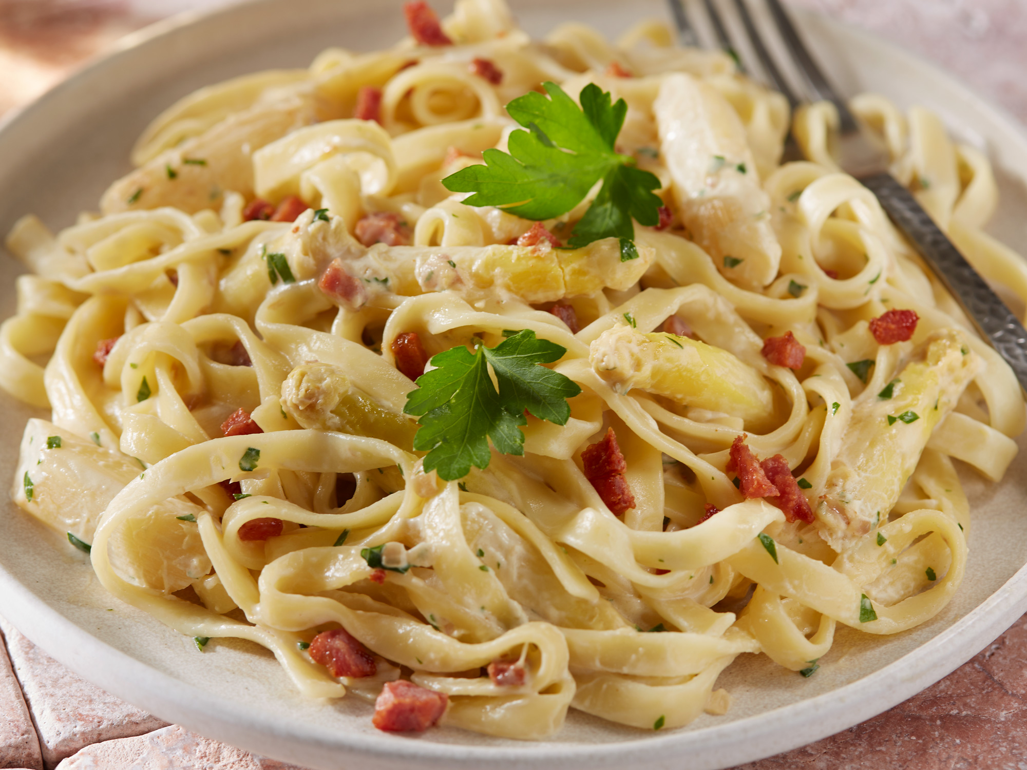 Nahaufnahme von Tagliatelle mit Sahnesauce, weißem Spargel und gebratenem Speck, garniert mit frischer Petersilie.